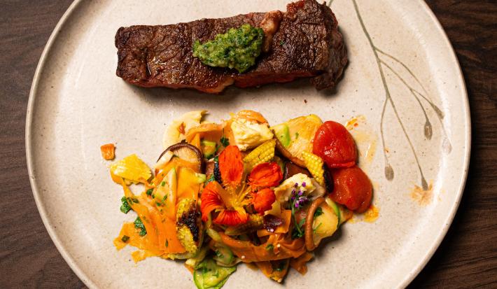 Fotografia vista de cima de um bife ancho com pesto de cerefólio e salada de cogumelos ao lado. A receita está em um prato grande, branco e raso, o qual está sobre uma mesa de madeira.