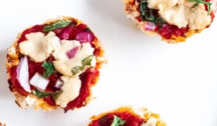 Fotografia em tons de vermelho em uma mesa branca, um prato redondo branco e vários pedaços pequenos de pizza feitas com massa de couve-flor e recheada de molho de tomate, rúcula e queijo mozarela.