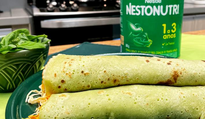 Fotografia em tons de verde com um prato verde ao centro. Em cima do prato existe algumas panquecas de cor esverdeada recheadas com frango desfiado e cenoura. Ao fundo existe uma embalagem de NESTONUTRI
