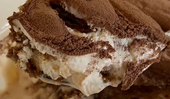 Fotografia em tons claros de um pedaço de tiramisù carregado por uma colher. No fundo tem o restante da receita em um recipiente refratário de vidro retangular.