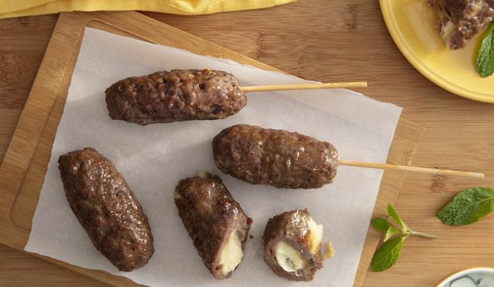 Foto da receita de Kafta Recheada com queijo. Observa-se 4 kaftas: duas no espeto, uma na tábua e outra cortada ao meio, expondo o recheio cremoso de queijo. Estão sobre uma tábua de madeira e há hortelãs decorando a foto.