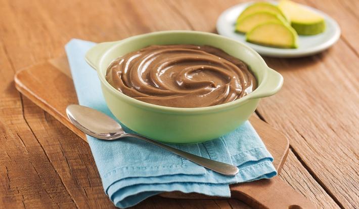 Fotografia em tons de marrom, em uma bancada de madeira de cor marrom. No centro, há uma tábua de madeira com um pano azul claro. Em cima, há um recipiente contendo o mousse com uma colher ao lado. Ao fundo, um pires contendo algumas fatias de abacate.
