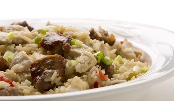 Fotografia em tons de bege, em uma bancada de madeira de cor branca. Ao centro, um prato rendo branco contendo o arroz com carne e palmito.
