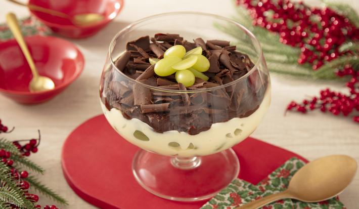 Fotografia mostra uma taça de sobremesa transparente com uma camada de creme branco com uvas. Por cima uma ganache de chocolate, com raspas de chocolate e pedaços de uva para decorar.