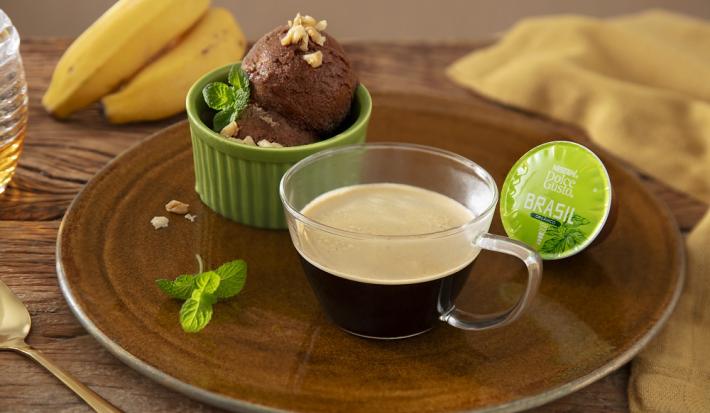 Foto em tons de marrom da receita de sorvete caseiro de banana e cacau servida em um pote de porcelana verde com uma xícara de dolce gusto brasil sobre uma tábua de madeira redonda em cima de uma mesa de madeira com bananas ao fundo