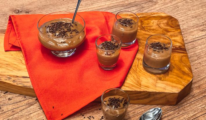 Fotografia em tons de laranja, em uma bancada de madeira, uma tábua de madeira, um pano laranja e 5 recipientes de vidro em cima dele, com os brigadeiros de colher com chocolate granulado em cima do pano e tábua.