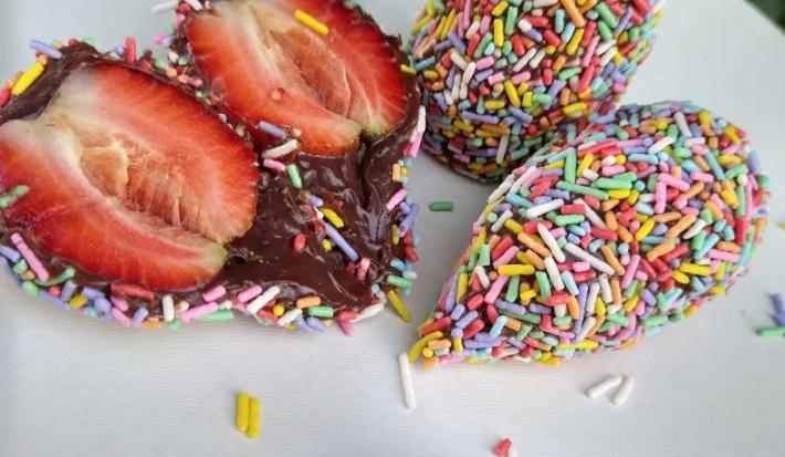Fotografia em tons coloridos em uma bancada de madeira branca. Ao centro, há um morango cortado ao meio e ao lado, há 2 coxinhas de brigadeiro e uma colher de brigadeiro.