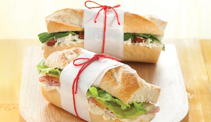 Fotografia em tons de amarelo em uma bancada de madeira com uma tábua de madeira com dois sanduíches na baguete com presunto e salada de alface.