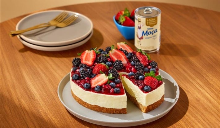 Fotografia mostra uma fatia de cheesecake de frutas vermelha sendo retirada e a lata de leite moça.