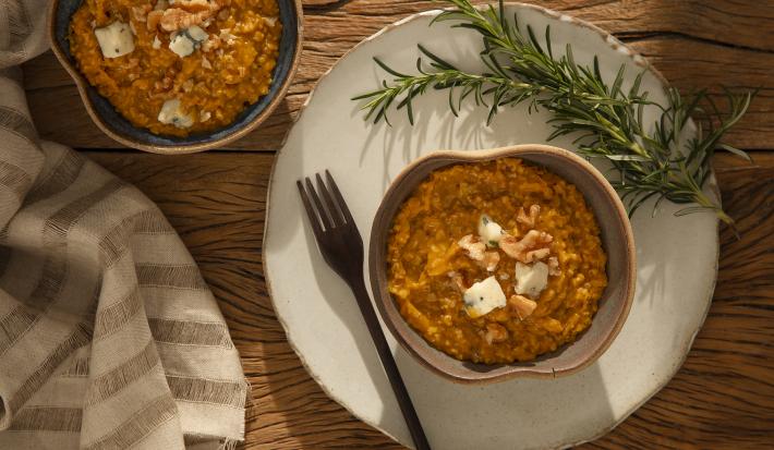 Foto em tons de laranja da receita de risoto de abóbora com gorgonzola servida em um bowl de cerâmica cinza sobre uma base de porcelana branca com um garfo ao lado além de um ramo de alecrim