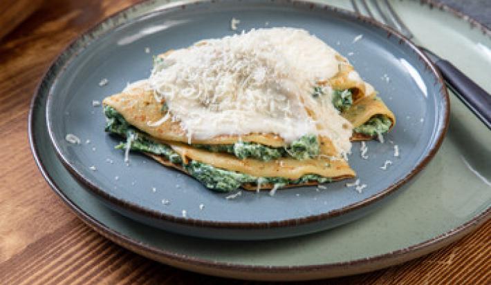 Fotografia de dois crepes de queijo e espinafre dobrados com molho branco por cima e queijo ralado. Os crepes estão em um prato azul raso, que está sore outro prato, um pouco maior, e ao lado, um garfo apoiado.