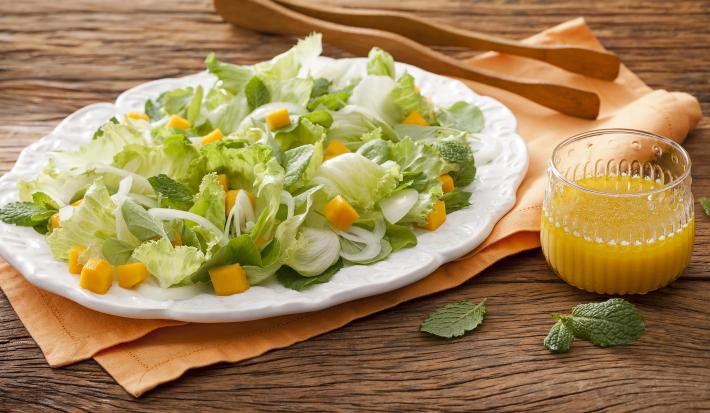 Fotografia em tons de marrom em uma bancada de madeira com um pano laranja e um prato branco oval com as hortaliças. Ao lado, um pegador de salada de madeira e um copo de vidro com suco de laranja.