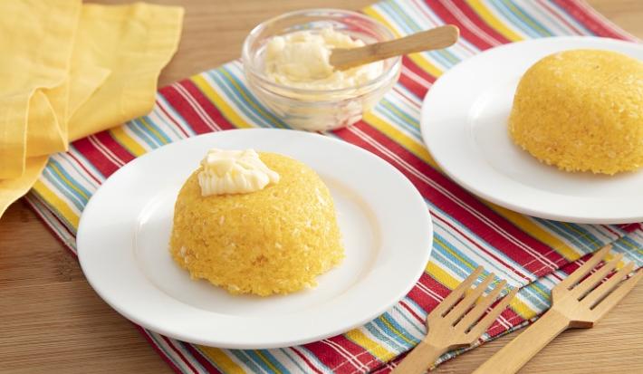 Foto em tons de amarelo da receita de cuscuz de micro-ondas com queijo servida em duas porções sobre pratos brancos de porcelana em cima de uma toalhinha colorida com garfos de madeira e um vidro com queijo ao lado