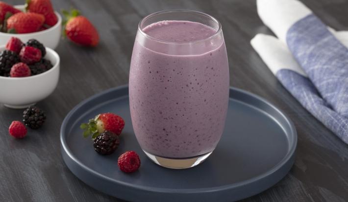Foto em tons de roxo da receita de shake de frutas vermelhas com chia servida em um corpo largo sobre uma base de porcelana azul escura circular com frutas vermelhas ao lado. Ao fundo, há dois recipientes com frutas vermelhas e um pano azul.
