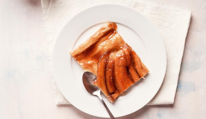 Fotografia vista de cima de uma torta de banana com bananas caramelizadas por cima em um prato de sobremesa branco, e uma colher ao lado. A sobremesa está em cima de um pano branco, que está sobre uma mesa branca.