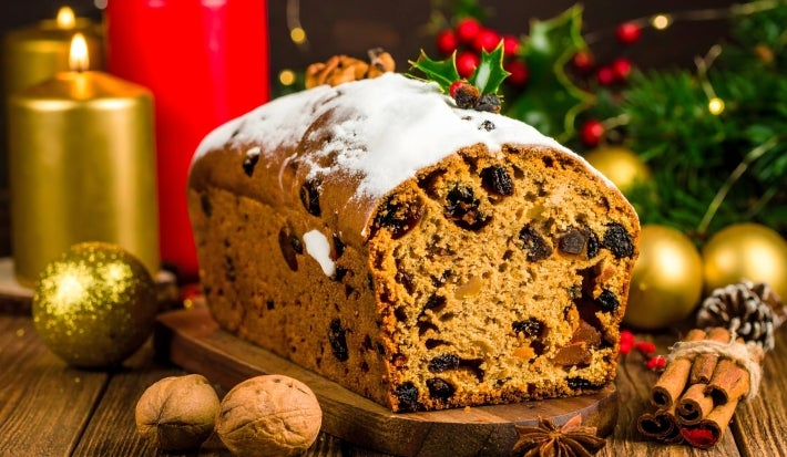 Fotografia de um bolo de frutas festivo, exibido sobre uma superfície de madeira, com textura densa e decorado com açúcar em pó. Ao redor, velas acesas, nozes e canela.