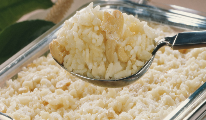 Fotografia em tons de branco e bege, ao centro colher de servir com arroz e cogumelos sobre refratário com o mesmo arroz, ao fundo folhas verdes como decoração.