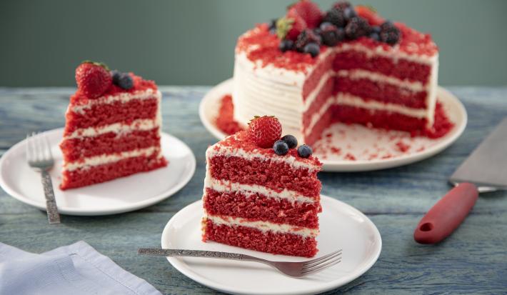 Imagem de dois pratos com fatias do bolo red velvet decorados com frutas vermelhas e ao fundo, o bolo inteiro.