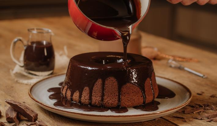 Fotografia em tons de vermelho em uma mesa de madeira com um prato branco com o bolo de chocolate em cima e uma calda de chocolate por cima. Uma panela vermelha em cima do bolo derramando a calda de chocolate.