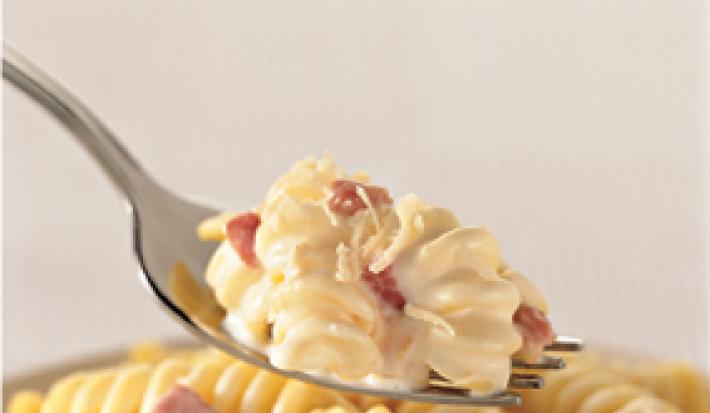 Fotografia em tons de amarelo de um garfo prateado com uma porção de macarrão parafuso cremoso com bacon sobre um pote com macarrão. Fotografia em tons de amarelo de um garfo prateado com uma porção de macarrão parafuso cremoso com bacon sobre um pote com macarrão.