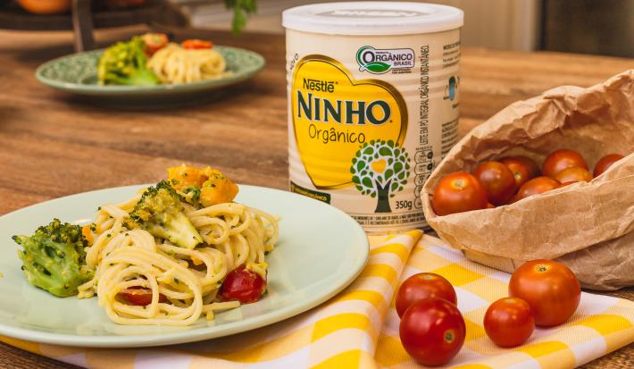 Fotografia em tons de amarelo de uma bancada de madeira com um pano xadrez amarelo e branco de um prato verde claro com o macarrão, brócolis, abóbora e tomate cereja. Ao fundo uma lata de NINHO.