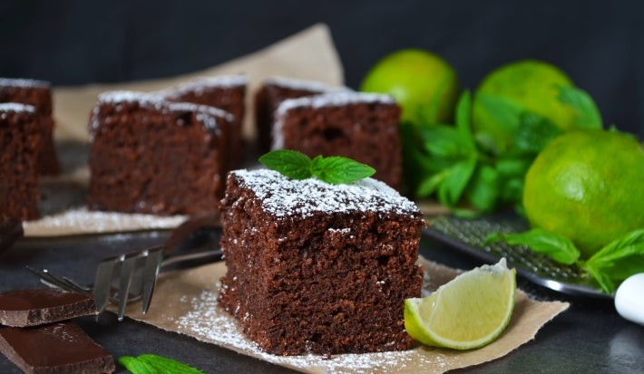 Pedaço de bolo de chocolate é o destaque, polvilhado com açúcar de confeiteiro e decorado com uma folha de hortelã. Ao lado, uma fatia de limão e mais bolos ao fundo, criando uma apresentação rústica e apetitosa.