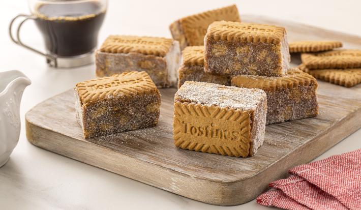 Fotografia de uma tábua de madeira com sete unidades de palha italiana com tostines especiais, ao lado de três biscoitos tostines. No fundo tem uma xícara de vidro com café.
