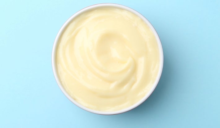 Fotografia em tons claros, vista de cima, de um recipeinte redondo, fundo, com o creme de abacaxi sobre uma mesa lisa azul clara.
