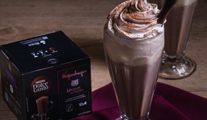 Fotografia em tons escuros de um copo de vidro de milkshake com uma bebida gelada de kopenhagen dole gusto com chantilly e uma língua de gato apoiado. O copo está ao lado da caixa dolce gusto.