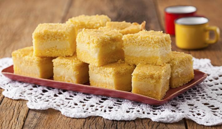 8 pedaços quadrados de bolo amarelo com uma lista branca no meio, dispostos em um prato vermelho que está em cima de uma toalha branca