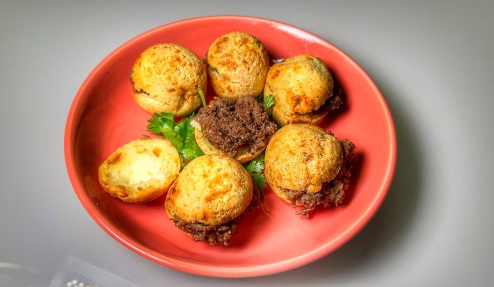 Fotografia de um prato vermelho sobre uma mesa branca. Dentro do prato tem seis pães de queijo recheados com carne de panela.