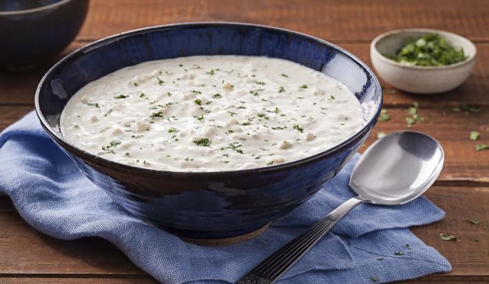 Fotografia em tons de marrom e azul de uma bancada de madeira, sobre ele um paninho azul com um recipiente azul com o creme de palmito e uma colher. Ao fundo um potinho redondo com salsinha.