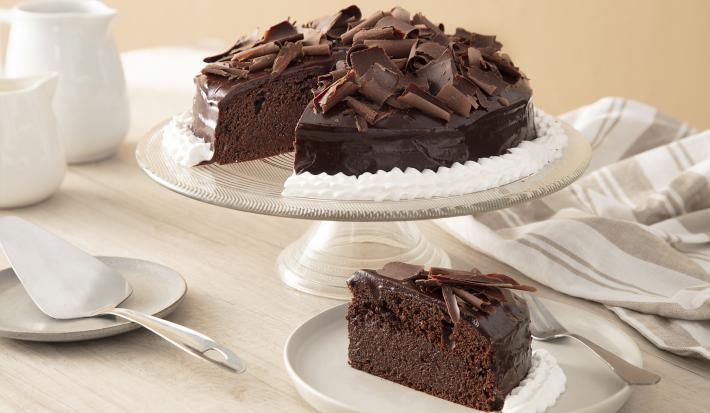 Foto da Receita de Bolo mousse de chocolate. Observa-se o bolo em uma boleira de vidro e, à frente, um prato de sobremesa com uma fatia do bolo. Louças de porcelana em tons off white decoram a foto e também uma espátula de prata