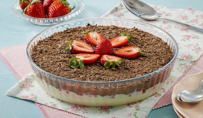 Imagem de uma travessa redonda de vidro com o bombom de travessa decorado com raspas de chocolate e morangos.