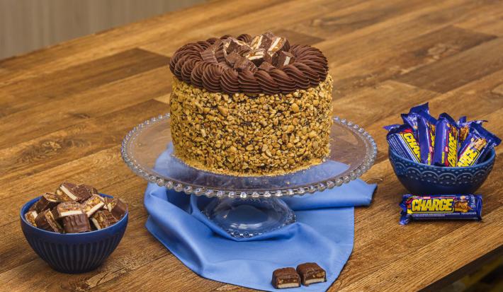 Foto aproximada da receita de Bolo Charge, decorado com amendoins e chocolate, sobre uma bancada de madeira, com alguns bombons Charge ao redor