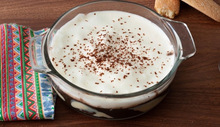 Em cima de uma mesa, recipiente redondo de vidro com creme de chocolate, decorado com chantilly e raspas de chocolate.