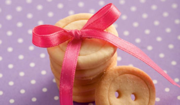 fotografia em tons de roxo, bege e rosa de uma bancada roxa com bolinhas brancas, contém biscoitinhos em formatos de botão com um laço rosa por cima.