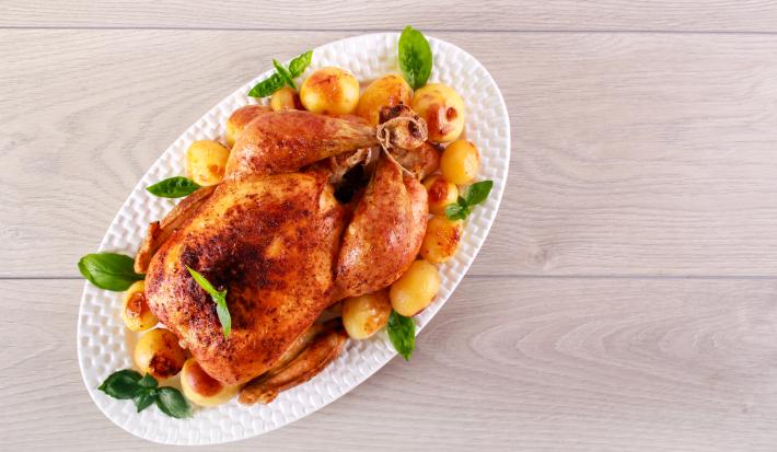 fotografia em tons de cinza, branco e bege de uma bancada cinza vista de cima. Contém um recipiente oval branco com frango assado e batatas bolinhas ao redor.