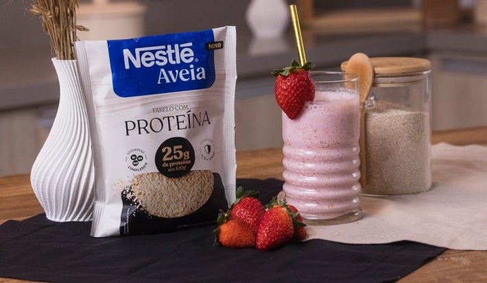 Smoothie pós-treino de morango em copo de vidro com canudo dourado e morango na borda, acompanhada por morangos frescos e pote de aveia