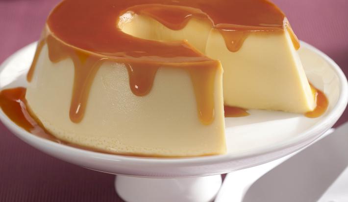Imagem de um prato com pé branco com o pudim de leite condensado e calda de caramelo cortado.