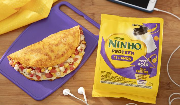 Fotografia em tons de roxo com um prato roxo ao centro. Em cima do prato existe uma receita de crepioca recheada com tomate, milho e palmito. Ao lado existe uma embalagem de Ninho Proteen.