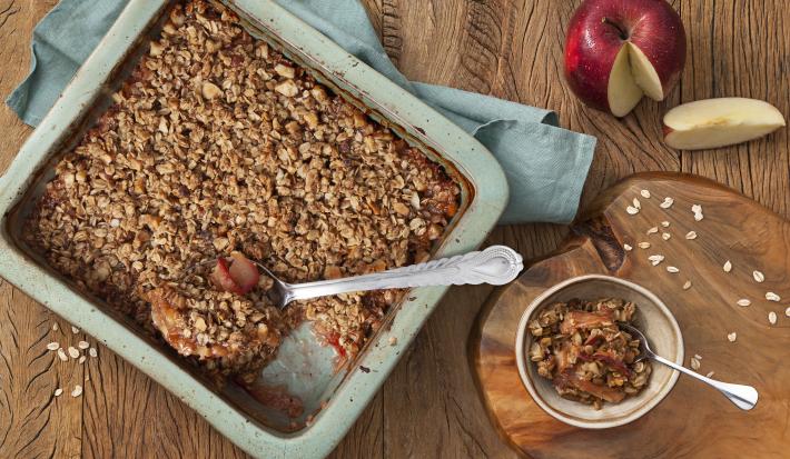 Fotografia em tons de azul em uma mesa de madeira escura com uma travessa retangular azul e o crumble de maçã e aveia dentro. Ao lado, uma tábua de madeira e um potinho com um pedaço do crumble. Ao fundo, uma maçã cortada ao meio.