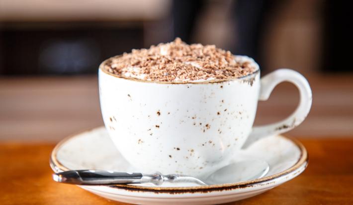 fotografia em tons de branco e marrom de uma bancada marrom vista de frente. Ao centro contém um pratinho redondo branco com uma xicara branca por cima, dentro da xicara contém a bebida e por cima raspas de chocolate, ao lado uma colher para mexer. fotografia em tons de branco e marrom de uma bancada marrom vista de frente. Ao centro contém um pratinho redondo branco com uma xicara branca por cima, dentro da xicara contém a bebida e por cima raspas de chocolate, ao lado uma colher para mexer.