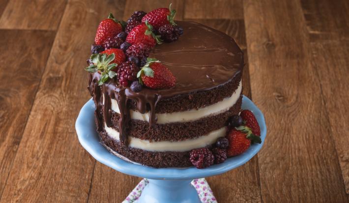 Imagem da receita de Naked Cake de Cacau, decorado em camadas marrons e brancas, com chocolate escorrendo, decorado com frutas vermelhas. O bolo está sobre um prato azul em uma bancada de madeira.