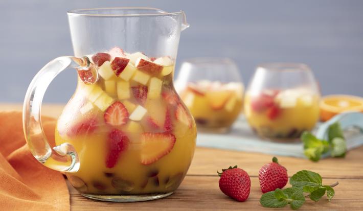 Imagem de uma jarra transparente com ponche e pedaços de frutas. Ao lado, morangos e folhas de hortelã e ao fundo dois copos da bebida.