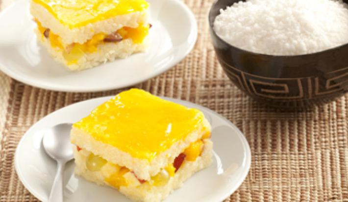 Fotografia em tons de amarelo em uma bancada de madeira com uma toalha de palha, dois pratos brancos redondos com um pedaço de pavê de tapioca em cada um. Ao lado, um potinho preto com coco ralado.