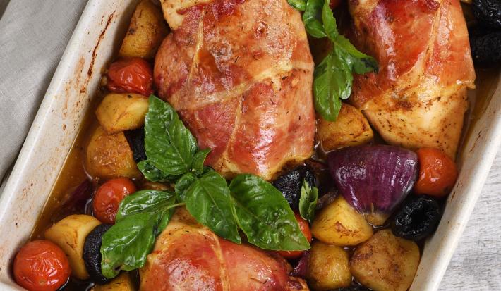 Fotografia vista de cima de três peitos de frango assados e recheados com maçã e ameixa no molho de laranja, e ao redor, vegetais assados que estão dentro de uma travessa de vidro na cor branca. O recipiente está sobre um pano cinza.