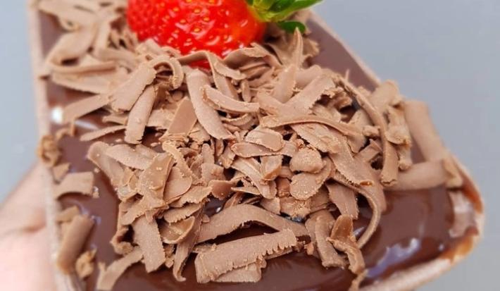 Fotografia de um bolo em forma retangular de chocolate com creme branco e brigadeiro. Por cima do bolo tem raspas de chocolate e morango.