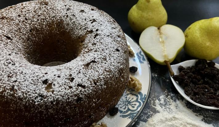 Foto da receita de bolo de pera com aveia em tons escuros servida em um prato de porcelana decorado sobre uma mesa com toalha adornada em azul e branco. Ao fundo, duas eras inteiras e uma metade como decoração, além de um recipiente com uvas passas.
