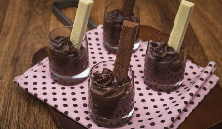 Fotografia em tons de rosa em uma bancada de madeira, uma tábua de madeira com um pano rosa e bolinhas pretas, quatro copinhos de vidro com brigadeiro de colher de chocolate dentro de cada um com um KitKat ao Leite e KitKat Branco dentro dos copinhos.